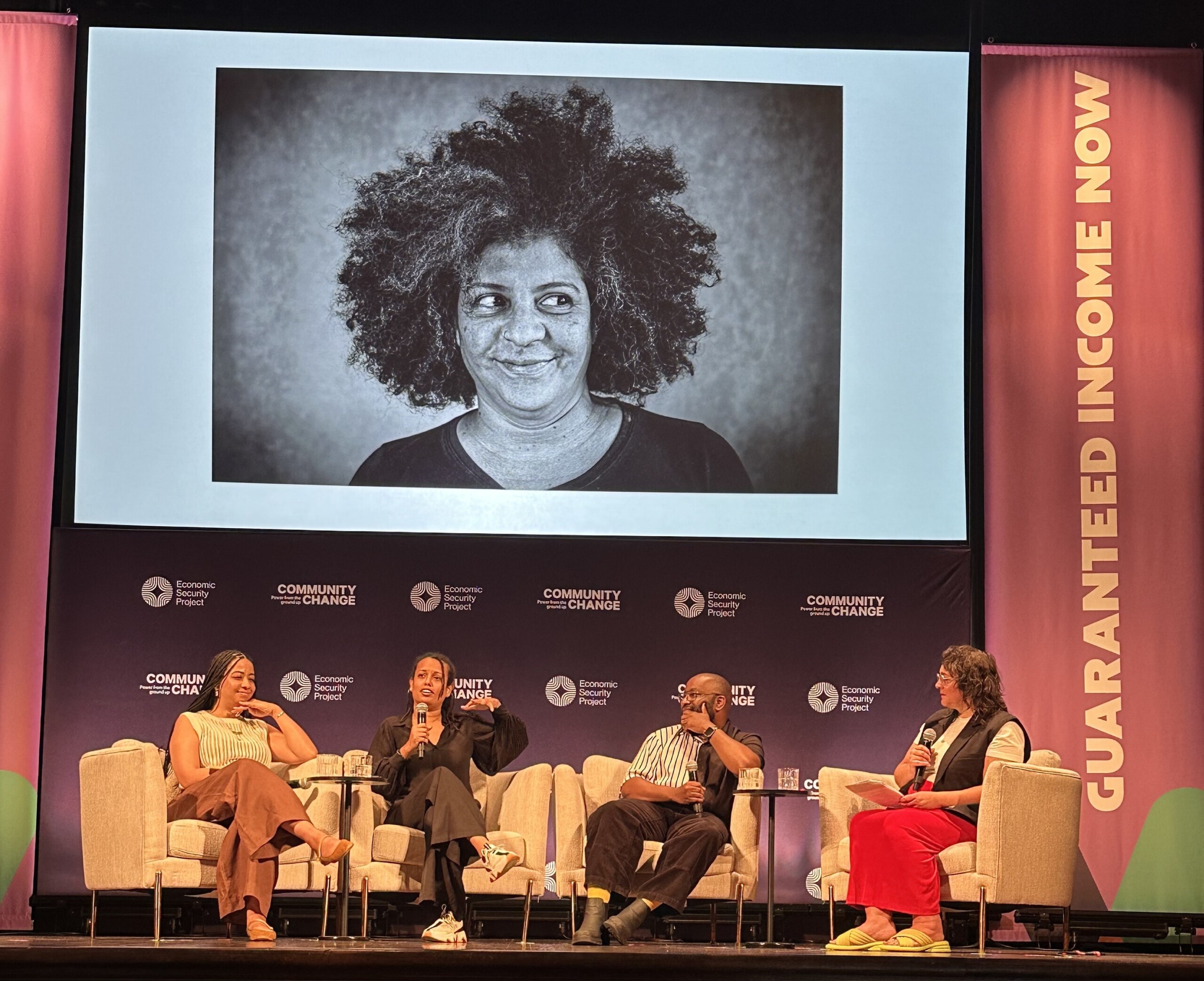 Maura Cuffie-Peterson, Ricardo Beaird, Claudia Maturell and moderator Cara Rose Defabio on stage at the Guaranteed Income Now conference with one of Claudia's photo portraits projected on the screen behind them.